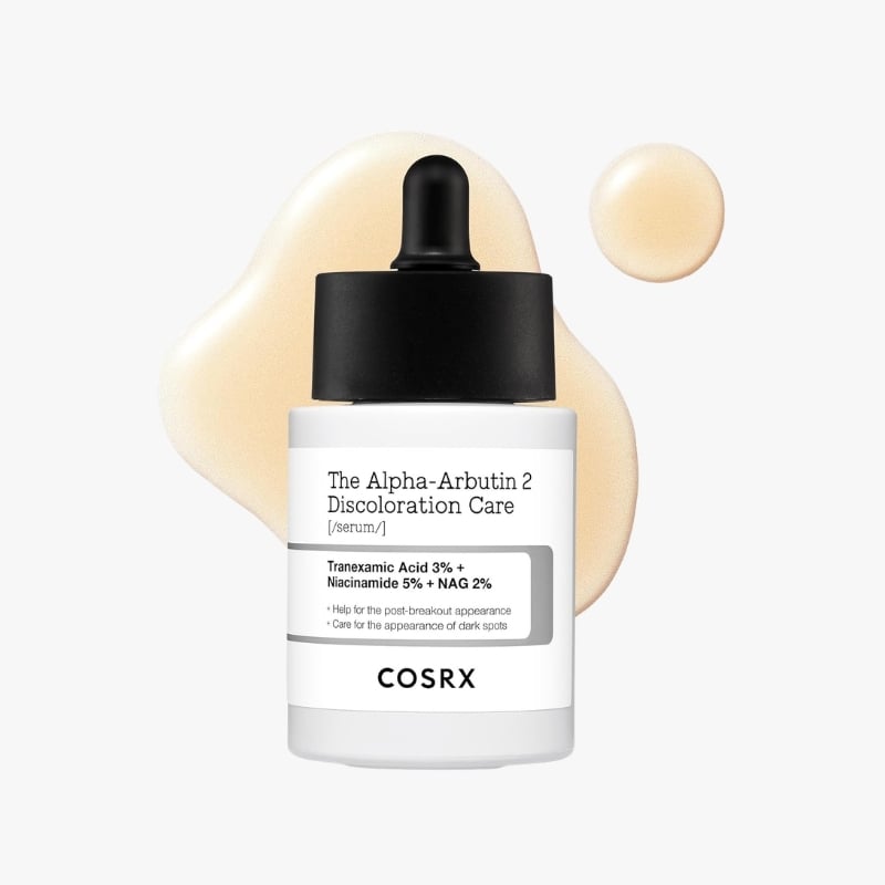 Bottle of COSRX The Alpha-Arbutin 2 Discoloration Care serum with dropper, label visible and beige serum splatter behind it