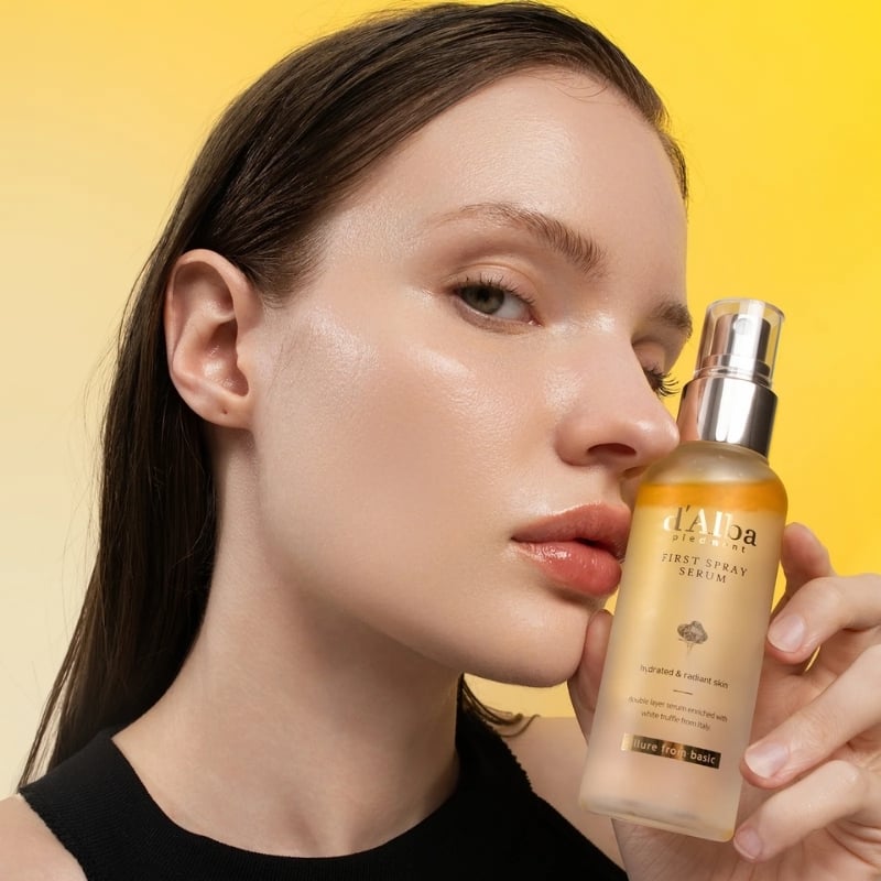 Woman holds a spray serum bottle near her face against a bright yellow background, showcasing the product.