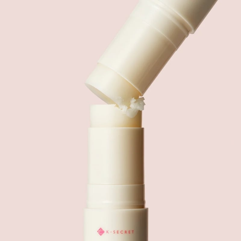 Cream lipstick tubes stacked vertically with one tilted tube shedding a small broken piece of product onto the gap, set against a pale pink background.