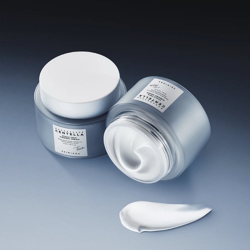 Two frosted glass jars of moisturizer with white lids, one open showing swirled white cream; the label reads 'Centella'.