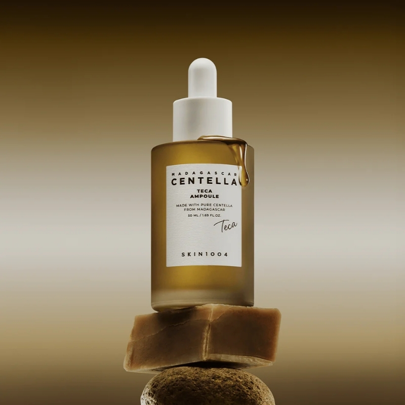 Brown glass dropper bottle labeled Centella Teca Ampoule sits on stacked rocks against a warm gradient background, skincare product scene.