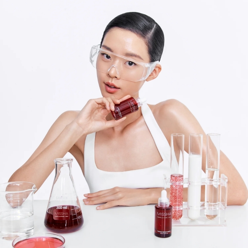 Young woman wearing safety goggles holds a small red bottle, surrounded by lab glassware and cosmetics on a white backdrop.