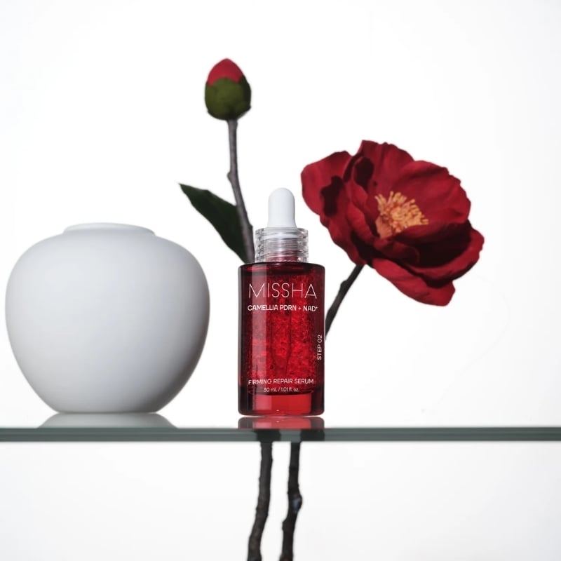 Missha firming repair serum bottle with dropper on a glass shelf, beside a white vase and red poppy in the background.