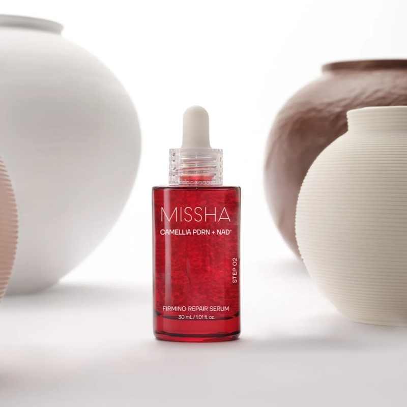 Red MISSHA serum bottle with dropper (Camellia PDRN + NAD+) on a white surface, flanked by white ceramic vases.