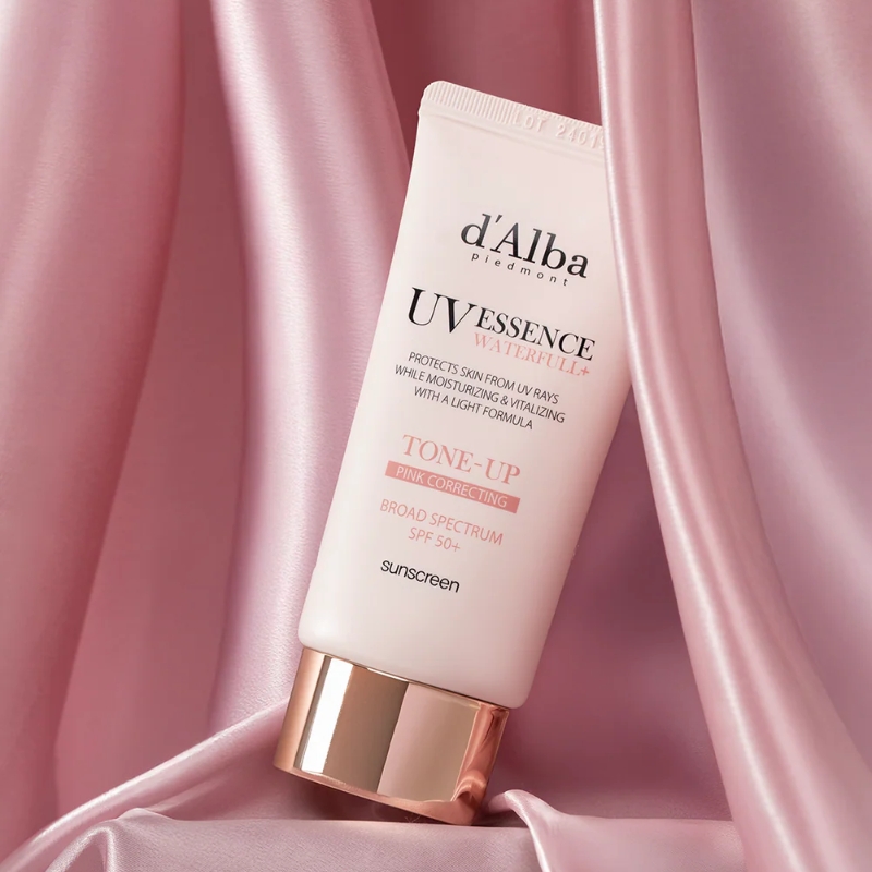 Tube of d'Alba UV Essence sunscreen on pink satin fabric; text on tube reads 'Tone-Up Pink Correcting' and 'SPF 50+'.