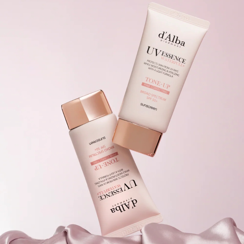 Two d'Alba UV Essence sunscreen tubes with rose-gold caps arranged diagonally on a soft pink satin backdrop, conveying a beauty product image.